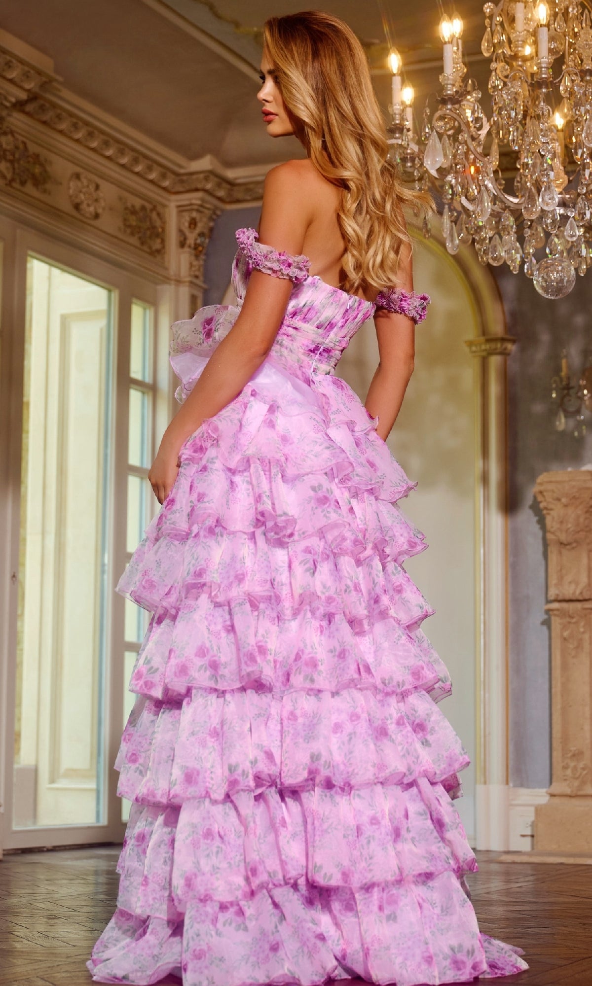 A woman models the Jovani 43786 long prom dress—a pink, ruffled, floral ball gown with an a-line skirt—standing indoors near a chandelier, facing slightly away from the camera.